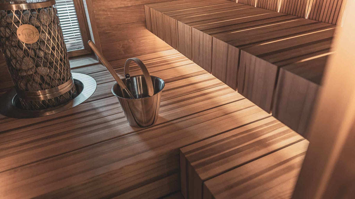 A sauna interior in warm wood with a stone-filled heater beside a window and a wooden bucket on the bench